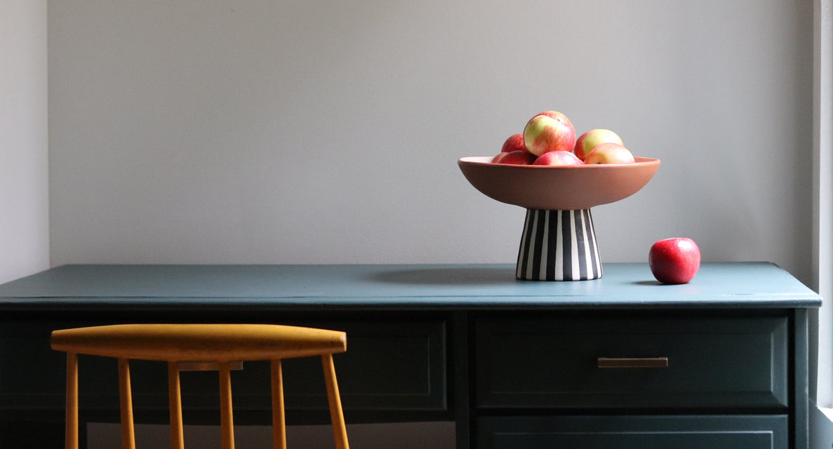 A dark teal writing desk with a black-and-white striped pedestal bowl filled with red apples and a single red apple on top, next to a mustard yellow wooden stool against a neutral gray wall.