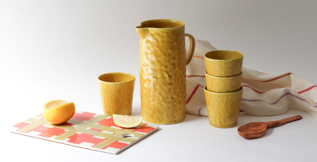 Yellow ceramic pitcher and cups on a white surface with a colorful mat and lemon slices.