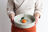 Person holding a ceramic bowl with a small orange fruit on a plain background