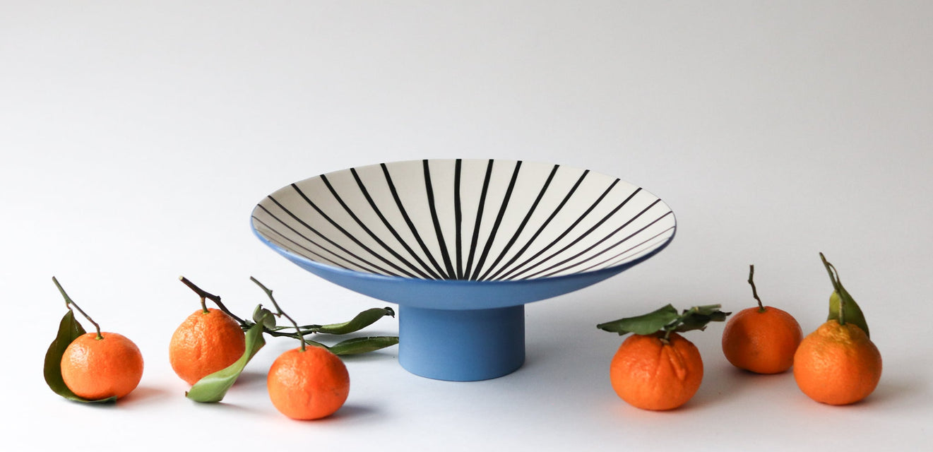 Decorative black and white striped bowl on a blue stand with oranges on a white background
